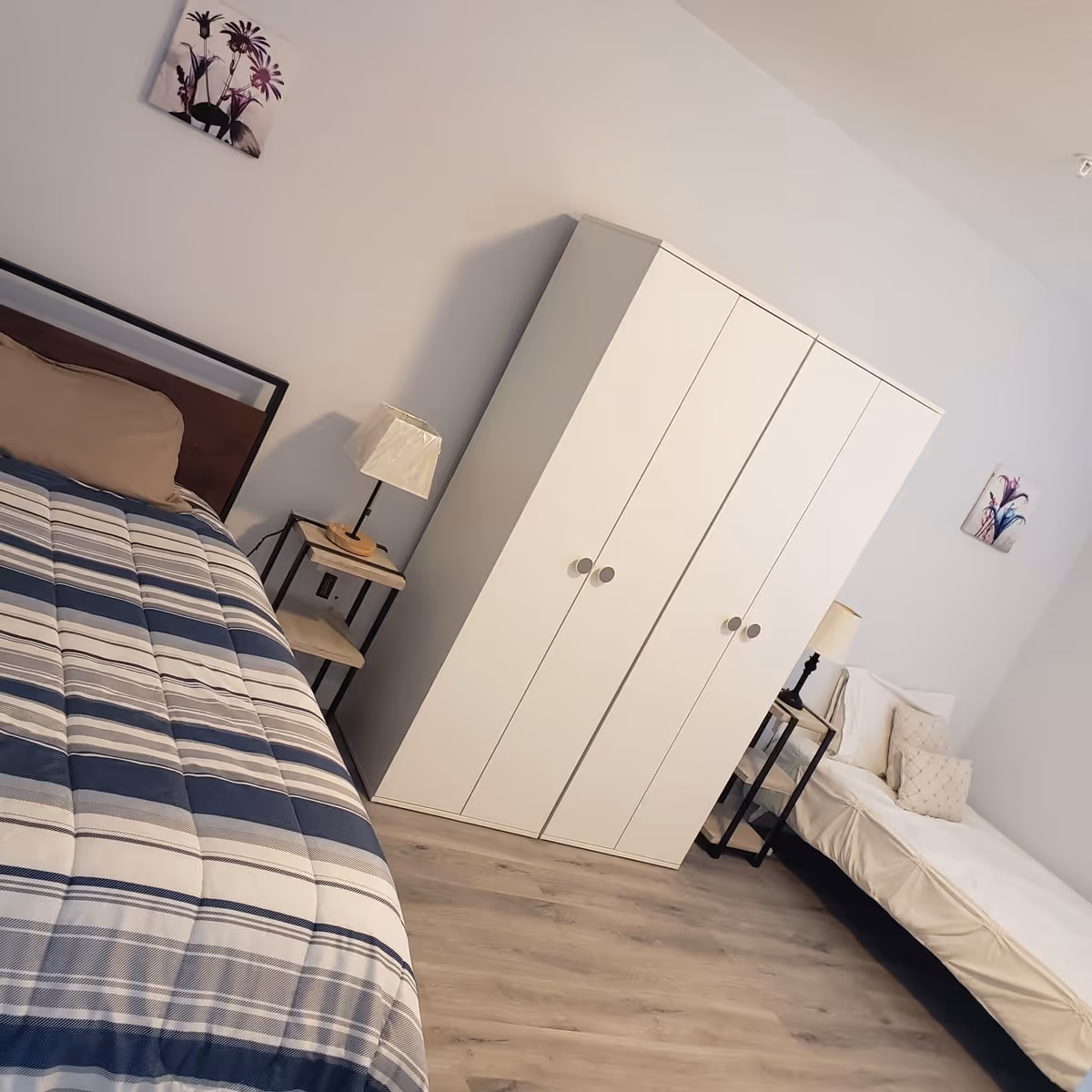 Two-bed bedroom with a striped comforter on the larger bed, a white wardrobe, bedside tables and lamps.