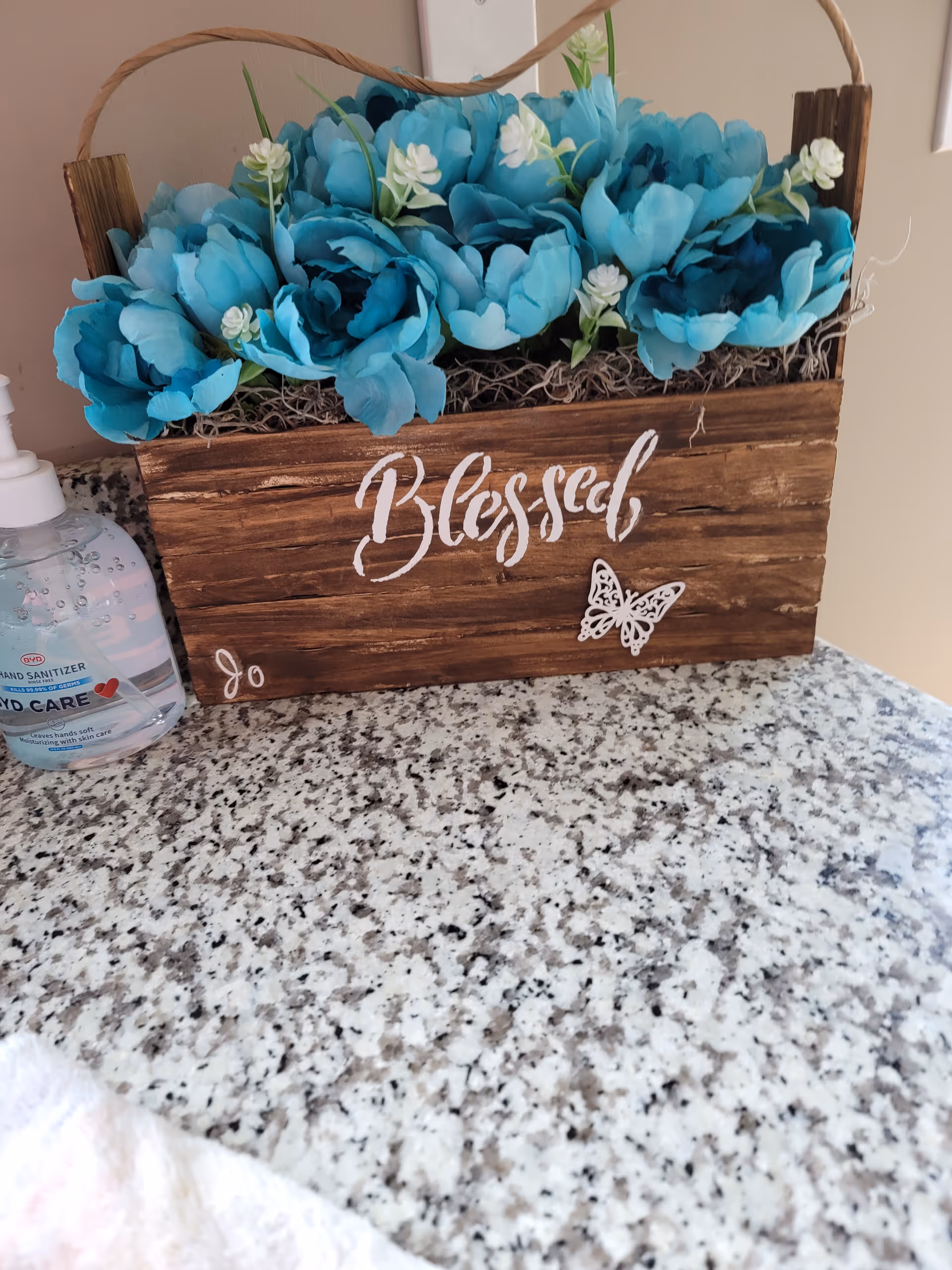 Wooden box planter labeled "Blessed" filled with blue artificial flowers sitting on a speckled countertop next to a hand sanitizer bottle.