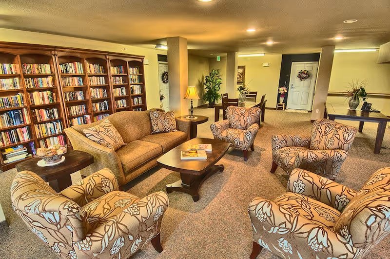 A cozy senior living facility common area with a brown sofa, four floral-patterned armchairs arranged around a wooden coffee table, and a large bookshelf filled with books along the wall. There are side tables with lamps and decorative plants, and a dining table with chairs in the background near the entrance door.