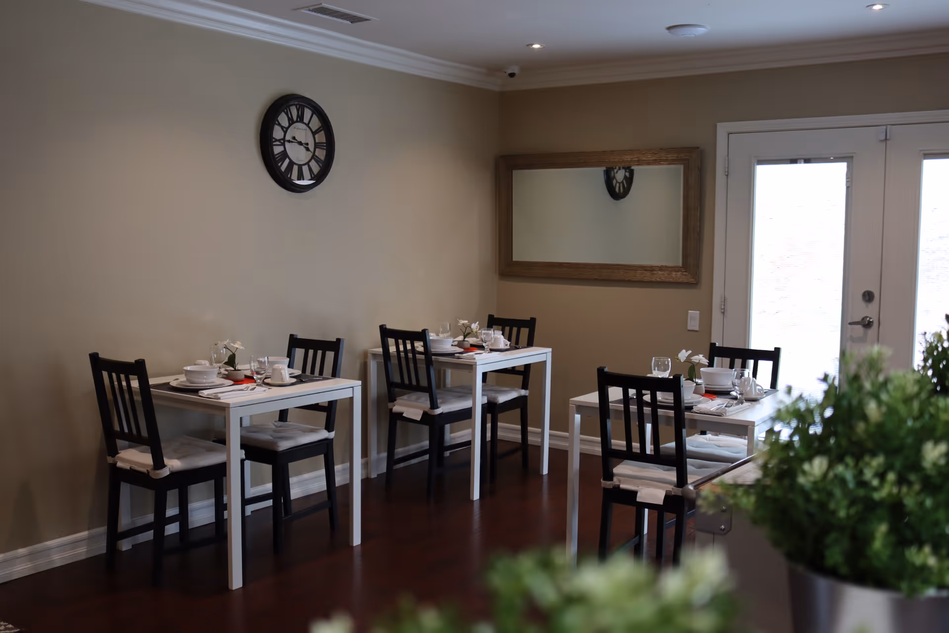 A small dining area with three white tables, each set with white dishes, cups, and small flower vases. The tables have black chairs with white cushions. A large wall clock and a rectangular mirror hang on the beige wall. There are double glass doors in the background and some green plants in the foreground.