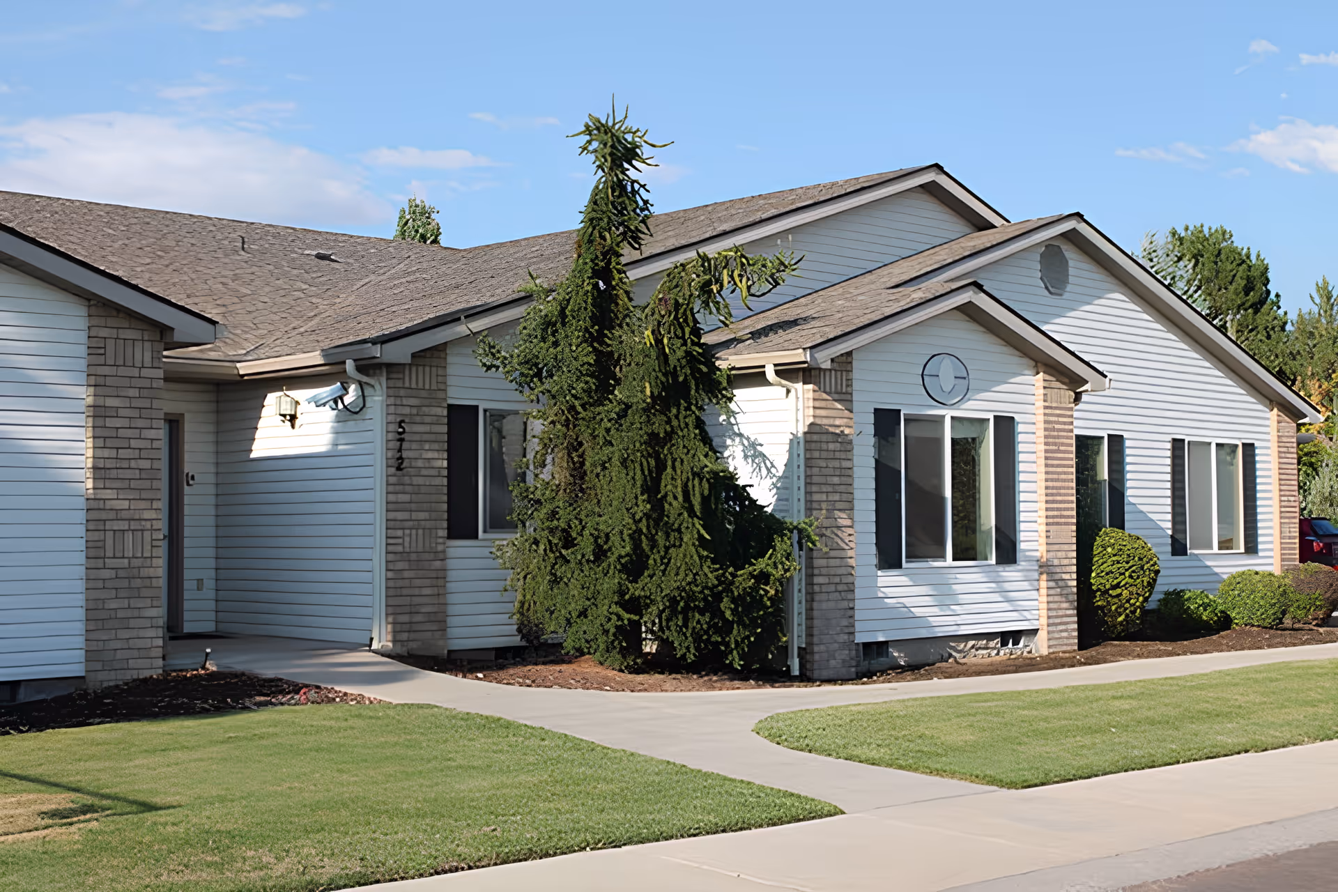 Exterior view of a single-story building with white siding and brick accents, surrounded by neatly trimmed bushes and a well-maintained lawn under a partly cloudy sky.