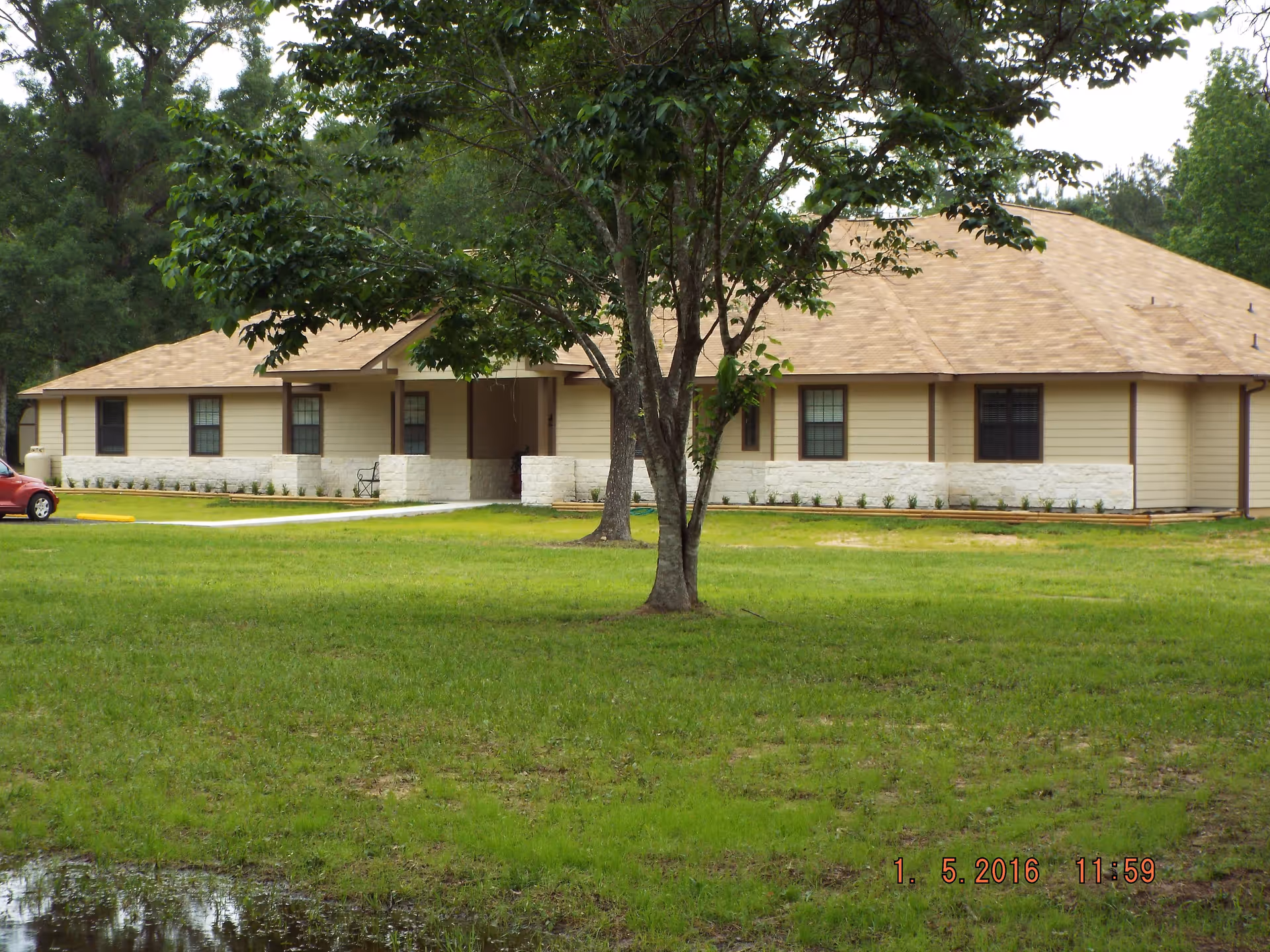 Single-story building with beige siding and a tan roof, surrounded by green grass and trees, with a red car parked on the left side.