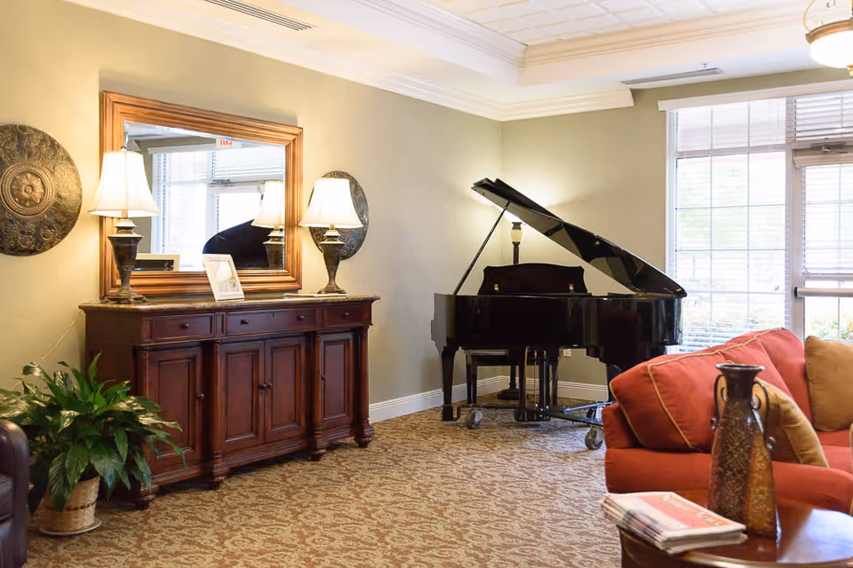 Bright communal living room with a grand piano, wooden sideboard topped by a mirror and lamps, and a red sofa by large windows.