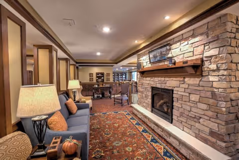 Cozy senior living room with a stone fireplace on the right, a blue sofa with orange pillows on the left, a patterned area rug on the floor, and a table lamp. In the background, there is a seating area with chairs and bookshelves.