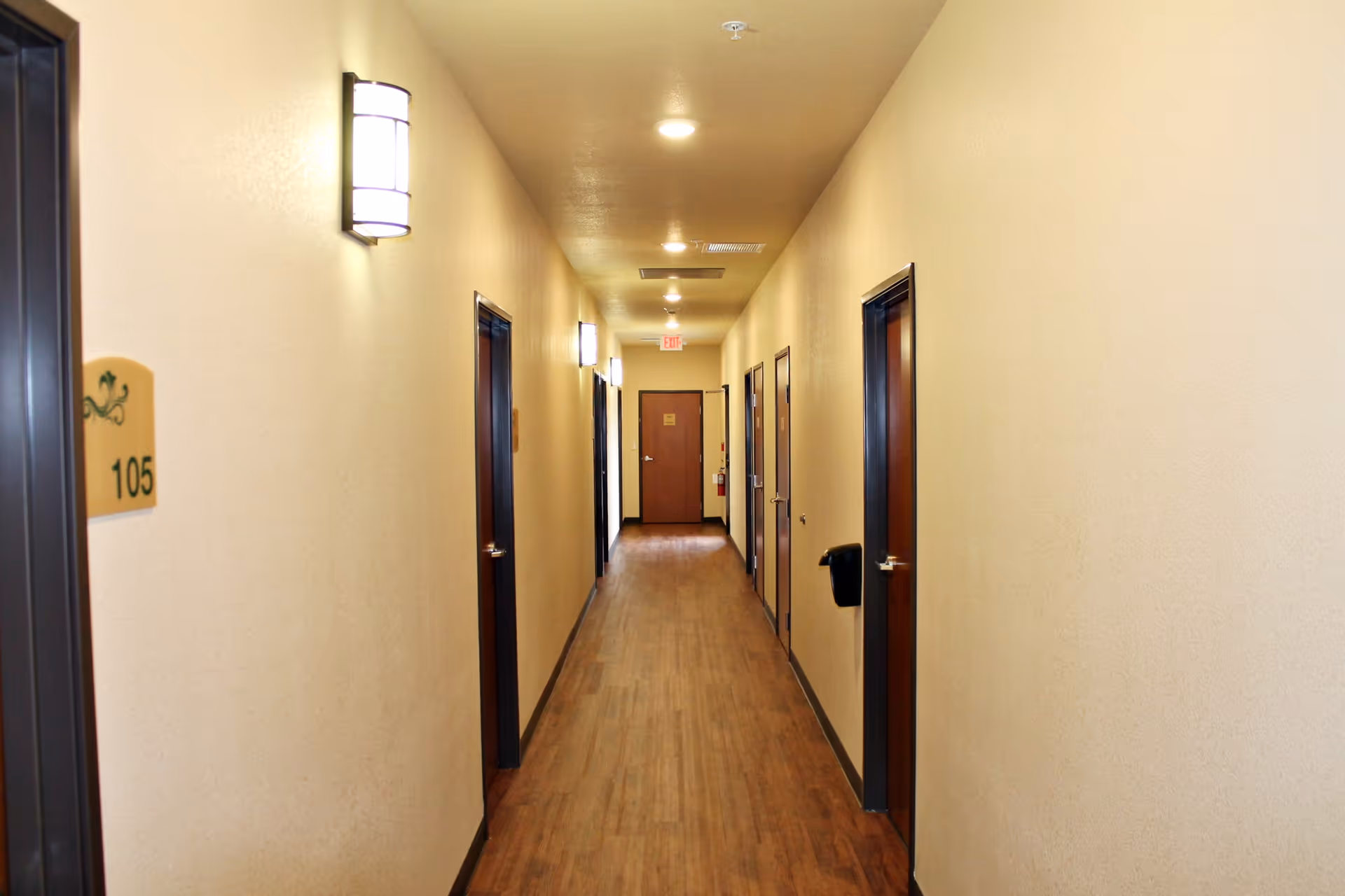 A long, narrow hallway in an assisted living facility with wooden flooring, beige walls, and several closed dark wooden doors on both sides. Wall-mounted lights illuminate the corridor, and a fire extinguisher is visible near the end of the hallway.