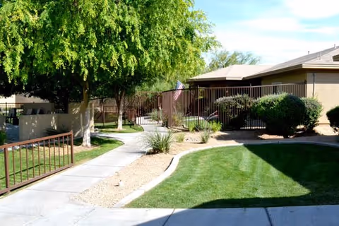 Sunlit courtyard with a concrete walkway, manicured lawns, shrubs, a tree, and a single-story building with a fenced patio and an American flag.