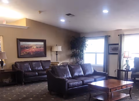 Sunlit lounge with leather sofas, a wooden coffee table, lamps, framed art, and windows.