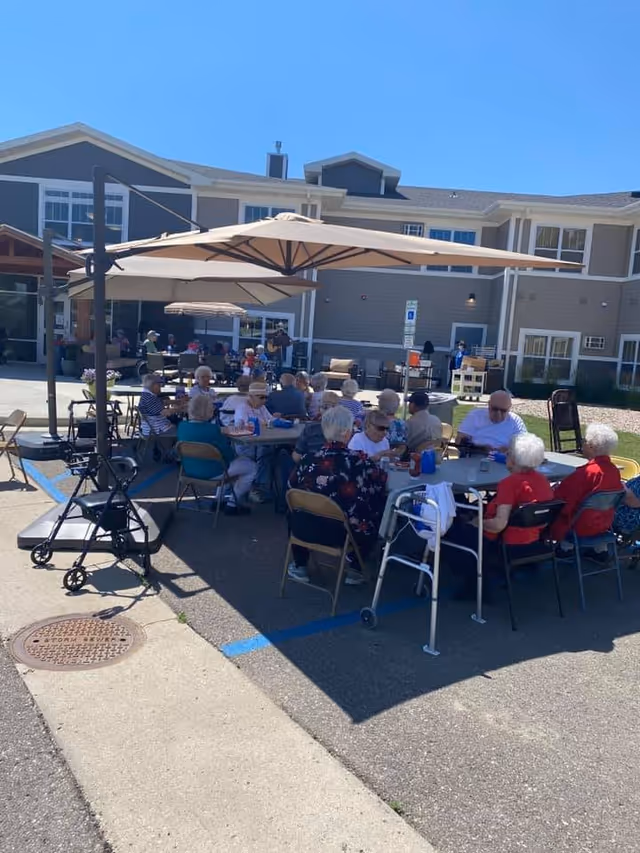 A group of elderly people sitting around tables outdoors under large umbrellas, socializing and enjoying a sunny day in front of a senior living facility building.