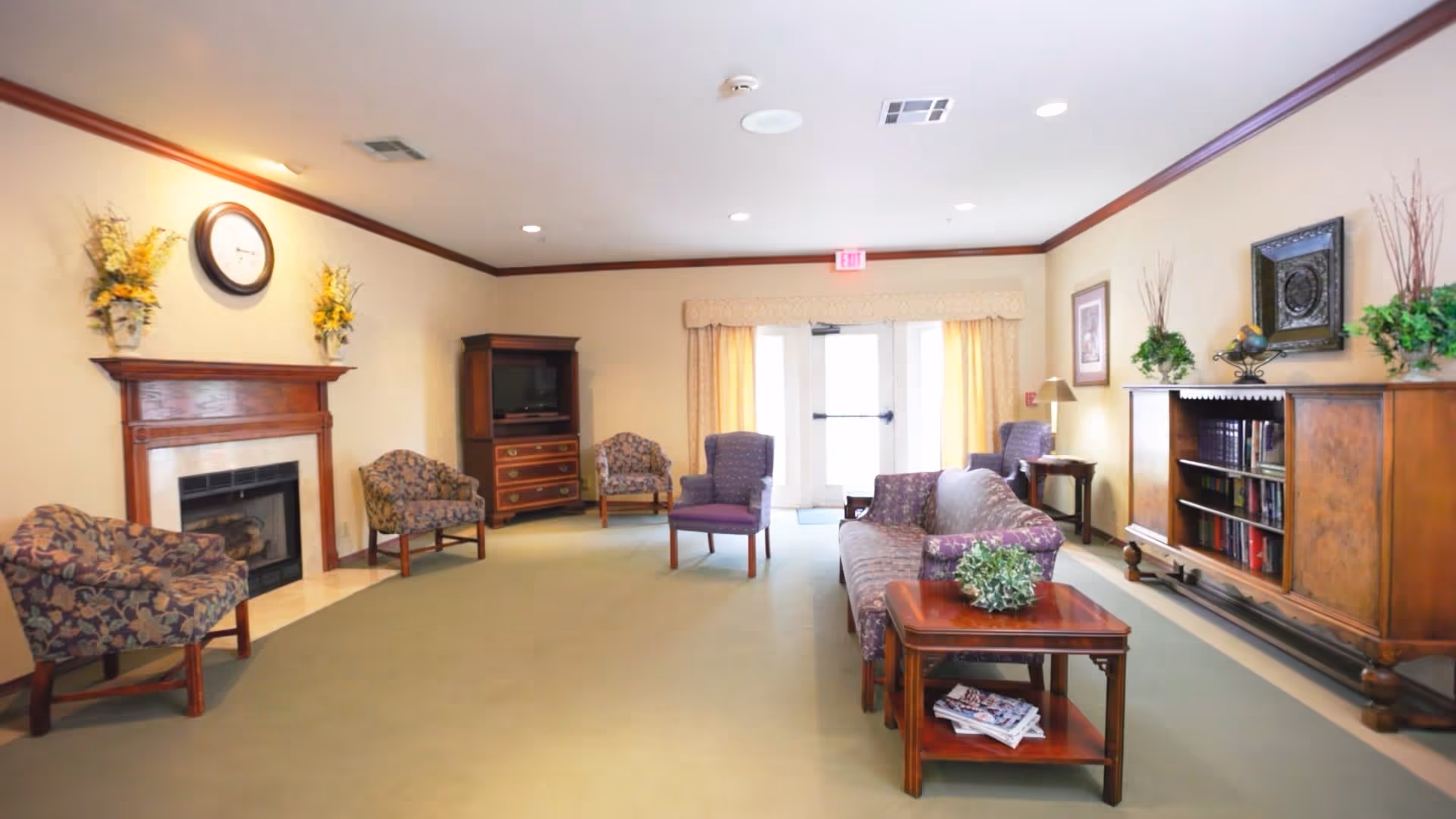 A cozy living room area in a senior living facility with floral upholstered chairs and a sofa, a wooden coffee table with a plant and magazines, a wooden fireplace with a clock and flower arrangements above it, a TV cabinet, and a wooden bookshelf with decorative plants and framed artwork on the walls. The room has beige walls, a green carpet, and a glass door with curtains letting in natural light.