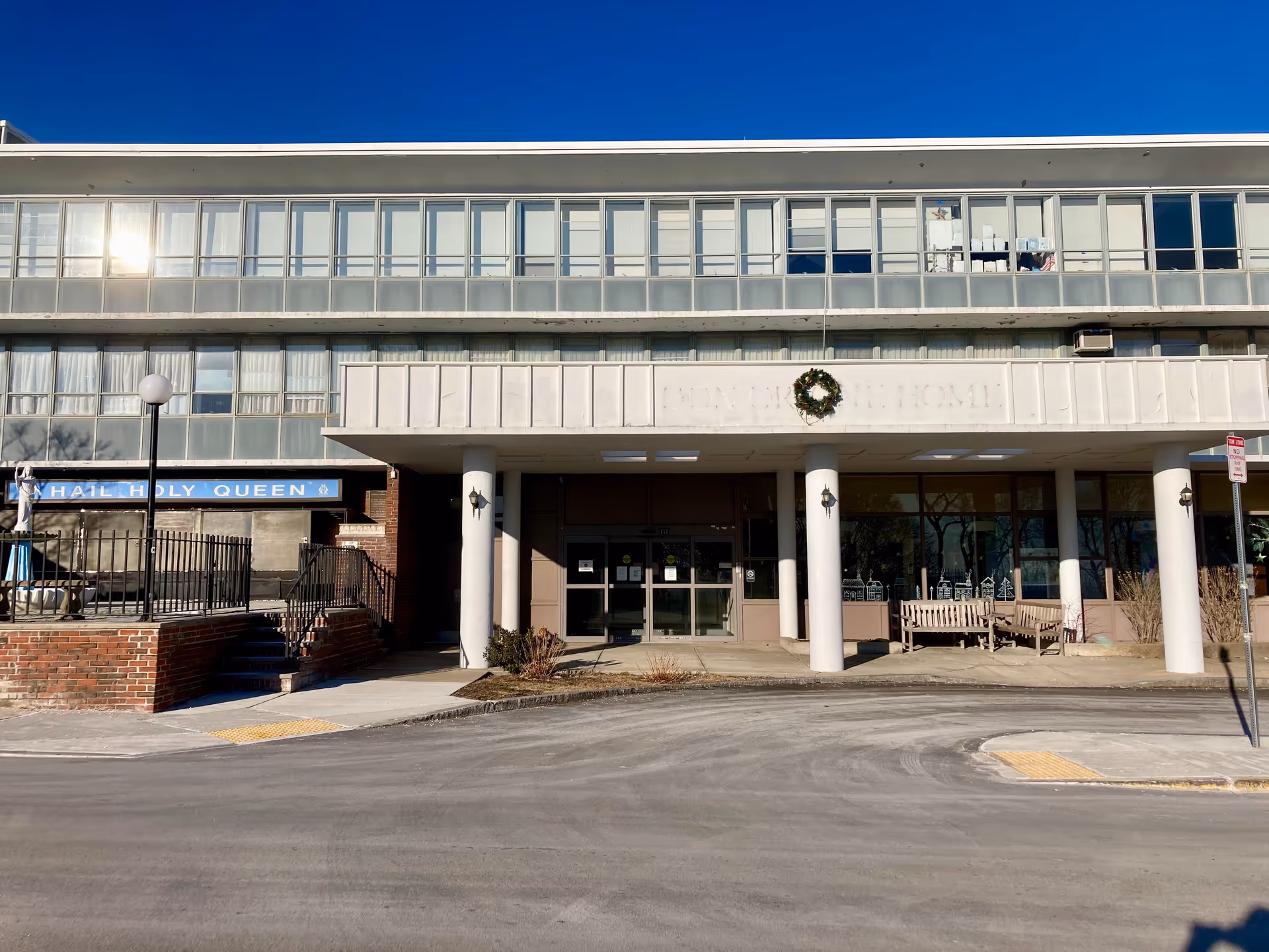 Front exterior view of a multi-story building with large windows and a covered entrance supported by white columns. There is a wreath hanging above the entrance and benches on the right side. A sign on the left reads 'HAIL HOLY QUEEN'. The sky is clear and blue.
