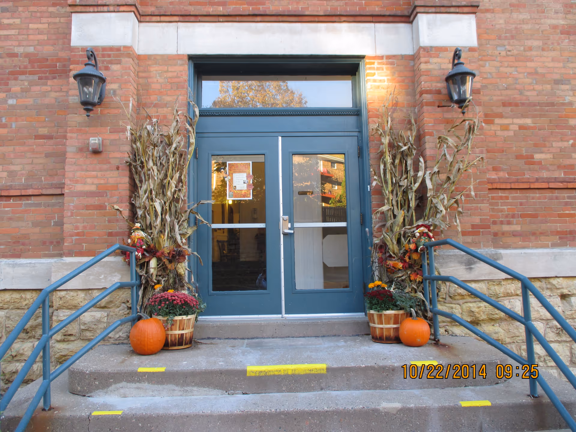 Brick building entrance with blue double doors flanked by pumpkins, potted flowers, and dried corn stalk decorations.