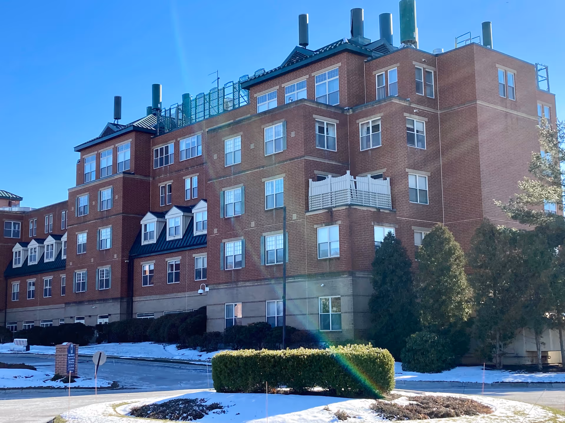 Red-brick multi-story senior living building with a circular driveway and patches of snow in front.
