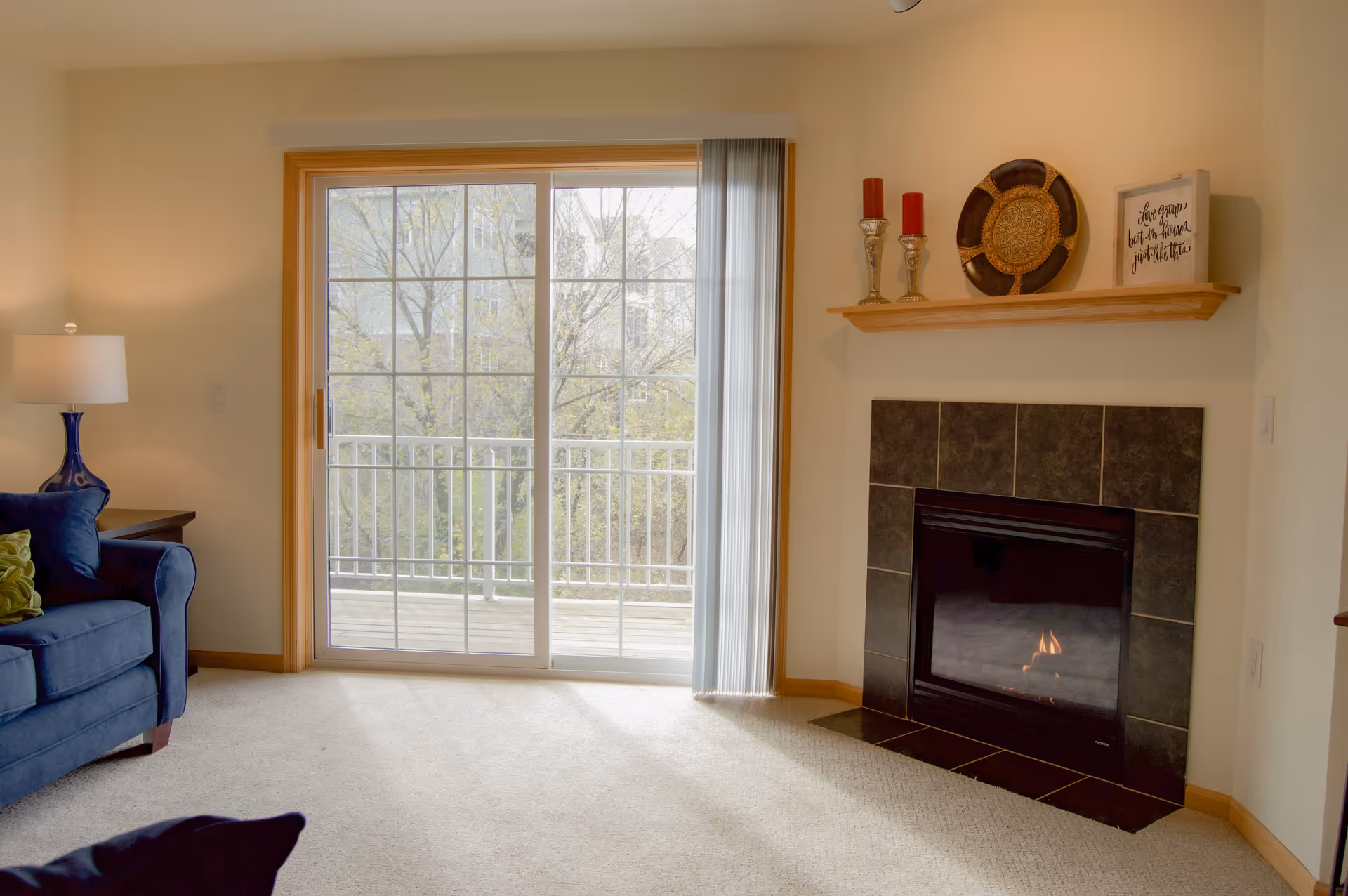 Cozy living room with a sliding glass door to a balcony, a tiled fireplace with mantel decor, and a blue sofa with a lamp.