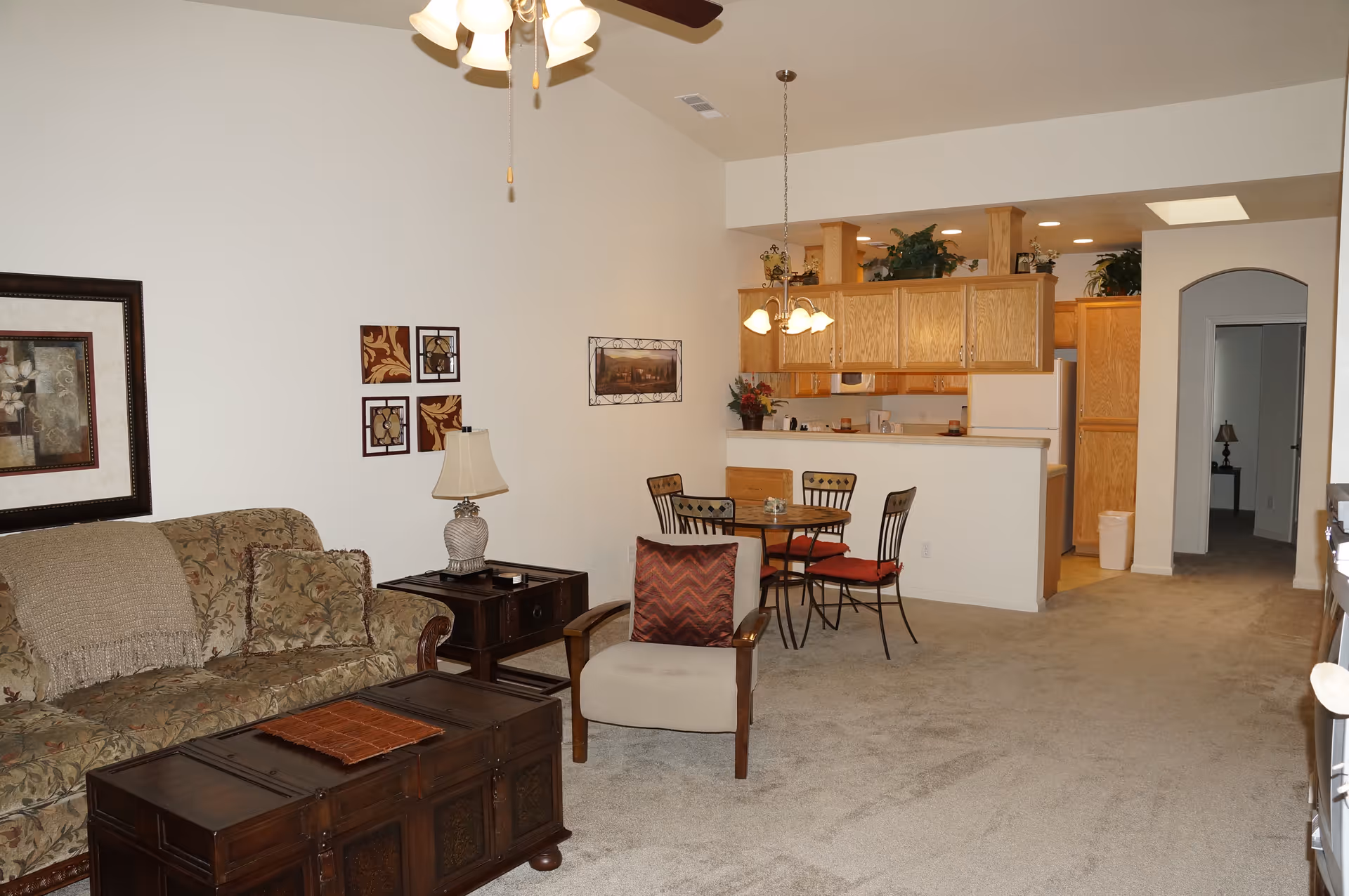 A living room and kitchen area in a senior living facility. The living room features a floral-patterned sofa with a beige throw, a wooden coffee table, a side table with a lamp, and an armchair with a red cushion. The kitchen has wooden cabinets, a white refrigerator, and a small dining table with four chairs. The walls are decorated with framed artwork, and the carpet is beige. Ceiling lights and a ceiling fan are visible.