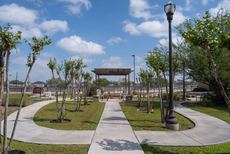 Outdoor garden area at The Heights of Alamo featuring a paved walkway that splits into three paths, young trees planted along the paths, a wooden pergola with benches underneath, a lamppost, and a partly cloudy blue sky.