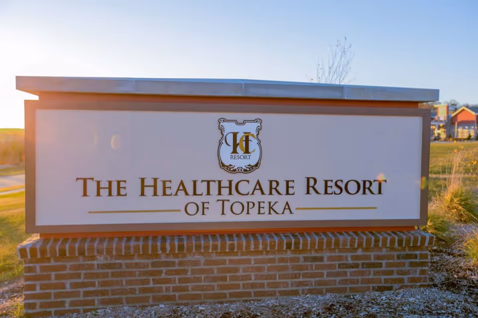 Outdoor brick-based sign reading "The Healthcare Resort of Topeka" with landscaping and buildings in the background.