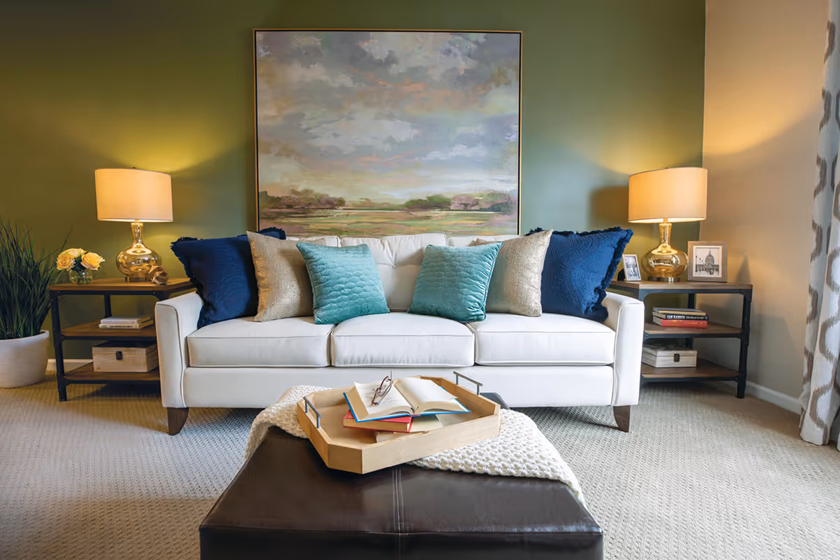 A cozy living room with a white sofa adorned with blue, teal, and beige pillows. On either side of the sofa are black side tables with gold lamps, books, and decorative items. A large landscape painting hangs on the green wall behind the sofa. In front of the sofa is a dark brown ottoman with a wooden tray holding an open book and reading glasses. The room has beige carpet and patterned curtains on the right side.