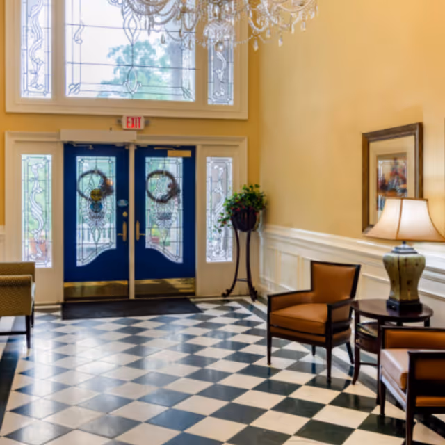A bright lobby with blue double entry doors, a large chandelier, black-and-white checkered floor, and seating with a side table and lamp.