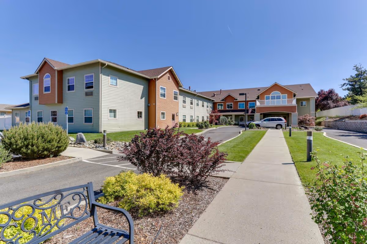 Exterior view of Siskiyou Springs Assisted Living Community building on a clear sunny day. The two-story building features light green and orange siding with multiple windows. A paved walkway leads to the entrance, flanked by well-maintained landscaping including bushes and small trees. A bench with the word 'WELCOME' is visible in the foreground.