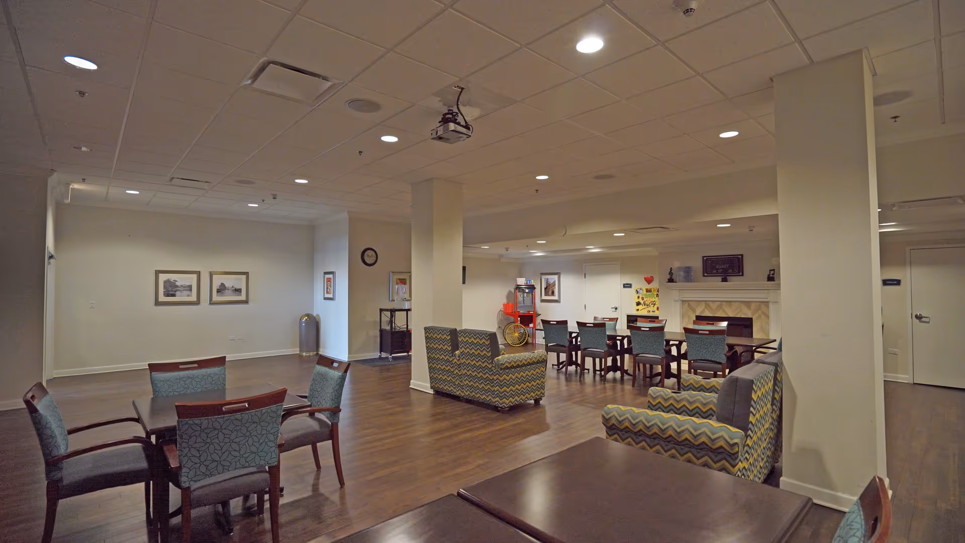 A spacious common area in a senior living facility with multiple seating arrangements including tables with chairs and patterned armchairs. The room has wooden flooring, a ceiling-mounted projector, framed pictures on the walls, and a fireplace in the background. The lighting is provided by recessed ceiling lights.