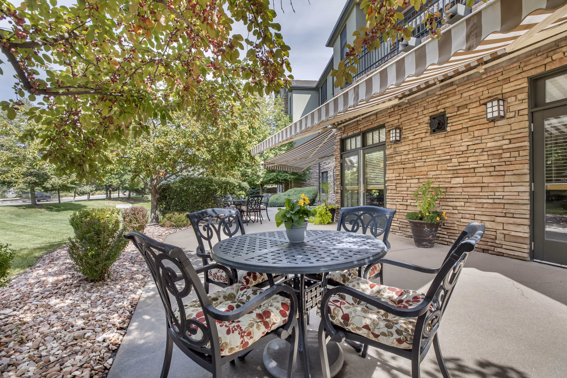 Outdoor patio with a round metal table and cushioned chairs under a striped awning beside a brick-clad building and landscaped lawn.