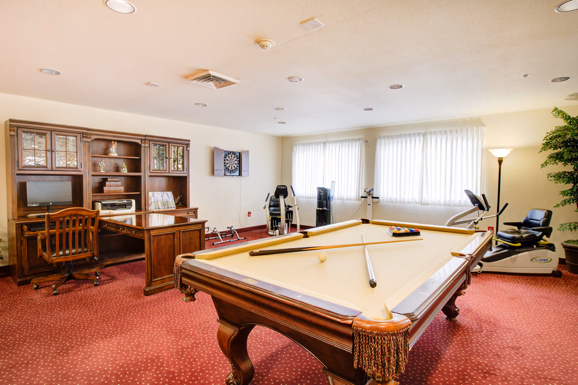 Well-lit interior activity room with a pool table in the foreground, exercise equipment, a dartboard, and a wooden desk and shelving along the back wall.