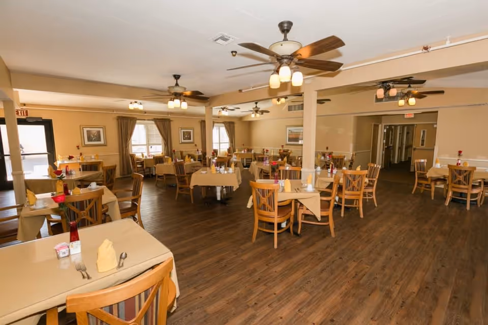A spacious dining room with multiple tables covered in beige tablecloths, each set with napkins, utensils, and small flower vases. The room has wooden chairs, ceiling fans with lights, framed artwork on the walls, and large windows with curtains allowing natural light to enter.