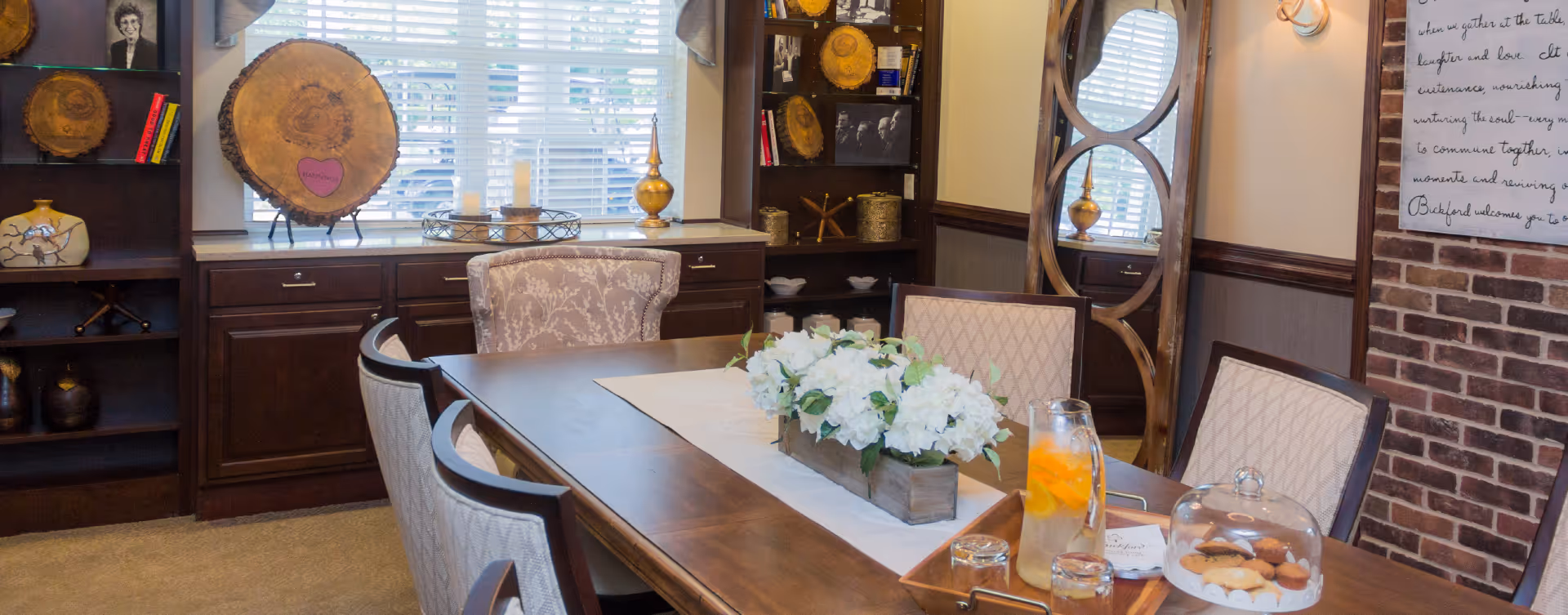 A cozy dining room with a wooden table set with a floral centerpiece, a glass pitcher with orange slices, glasses, and a tray of cookies under a glass dome. The room features upholstered chairs, built-in dark wood cabinetry with decorative items, a large window with blinds, a tall mirror with a wooden frame, and a brick accent wall with a framed quote.
