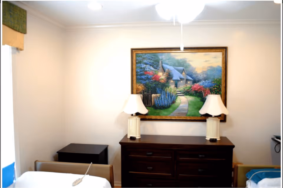 A bedroom in an assisted living facility featuring a wooden dresser with two white lamps on top, a colorful framed painting of a cottage with a garden hanging above the dresser, and parts of two beds visible on either side of the dresser.