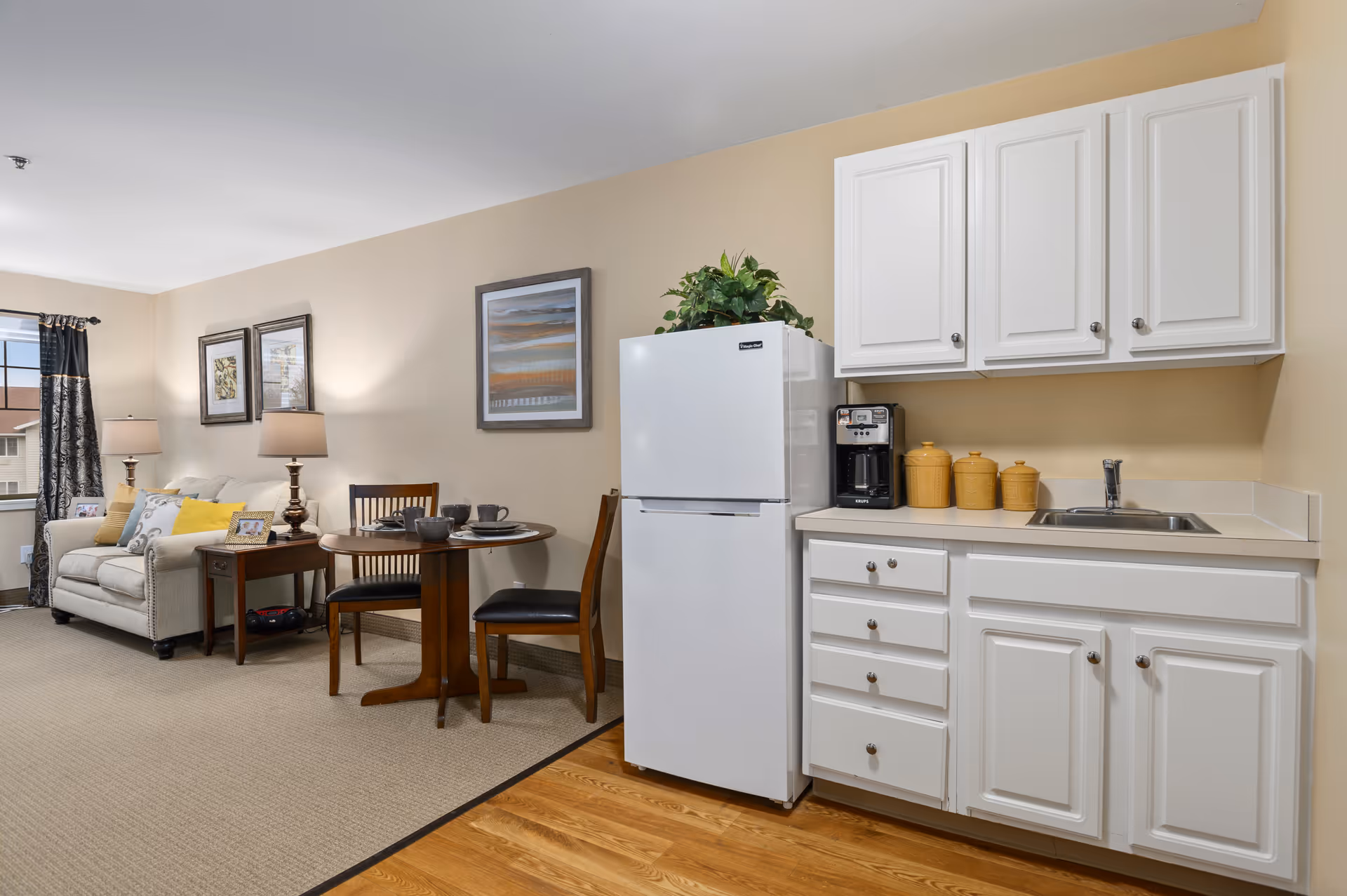 Open-plan senior living apartment showing a kitchenette with white cabinets and refrigerator beside a small dining table and a living area with a sofa and lamps.