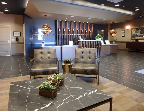 A modern reception area with a dark marble coffee table adorned with small potted plants in the foreground, two cushioned chairs behind the table, and a reception desk with a staff member in the background. The space features pendant lights, dark tiled flooring, and decorative wall elements.