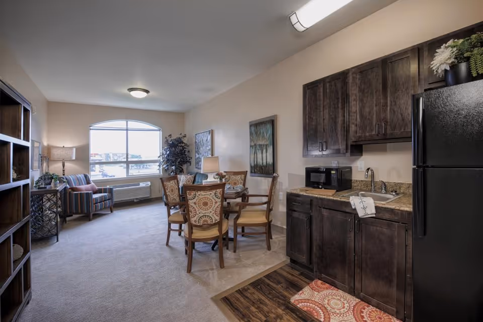 A bright senior apartment living area with a small kitchenette, round dining table and chairs, an armchair by a large window, and dark wood cabinets and refrigerator.