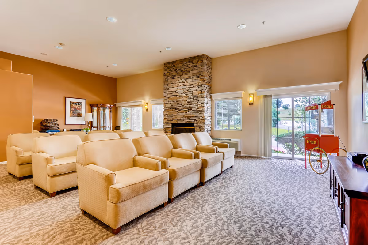 A spacious living room with beige armchairs arranged in rows facing a television mounted on the wall. The room features a stone fireplace, large windows with white blinds, and a popcorn machine near a sliding glass door that leads outside. The walls are painted in warm tones, and the carpet has a patterned design.