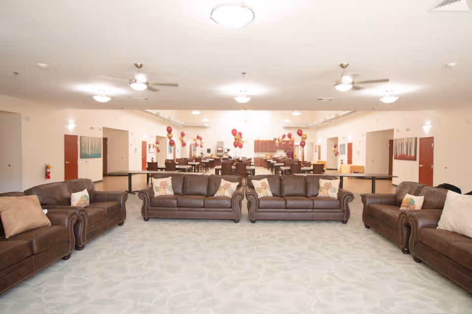 A spacious senior living common area with four brown leather sofas arranged in a square facing inward. The room has light-colored walls and carpeted flooring with a subtle pattern. In the background, there are several tables and chairs decorated with red and gold balloons, and a kitchen area with cabinets and appliances is visible. Ceiling fans and lights are mounted on the white ceiling.