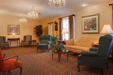 A warmly decorated sitting room with upholstered armchairs and a sofa, chandeliers, framed artwork, and potted plants.