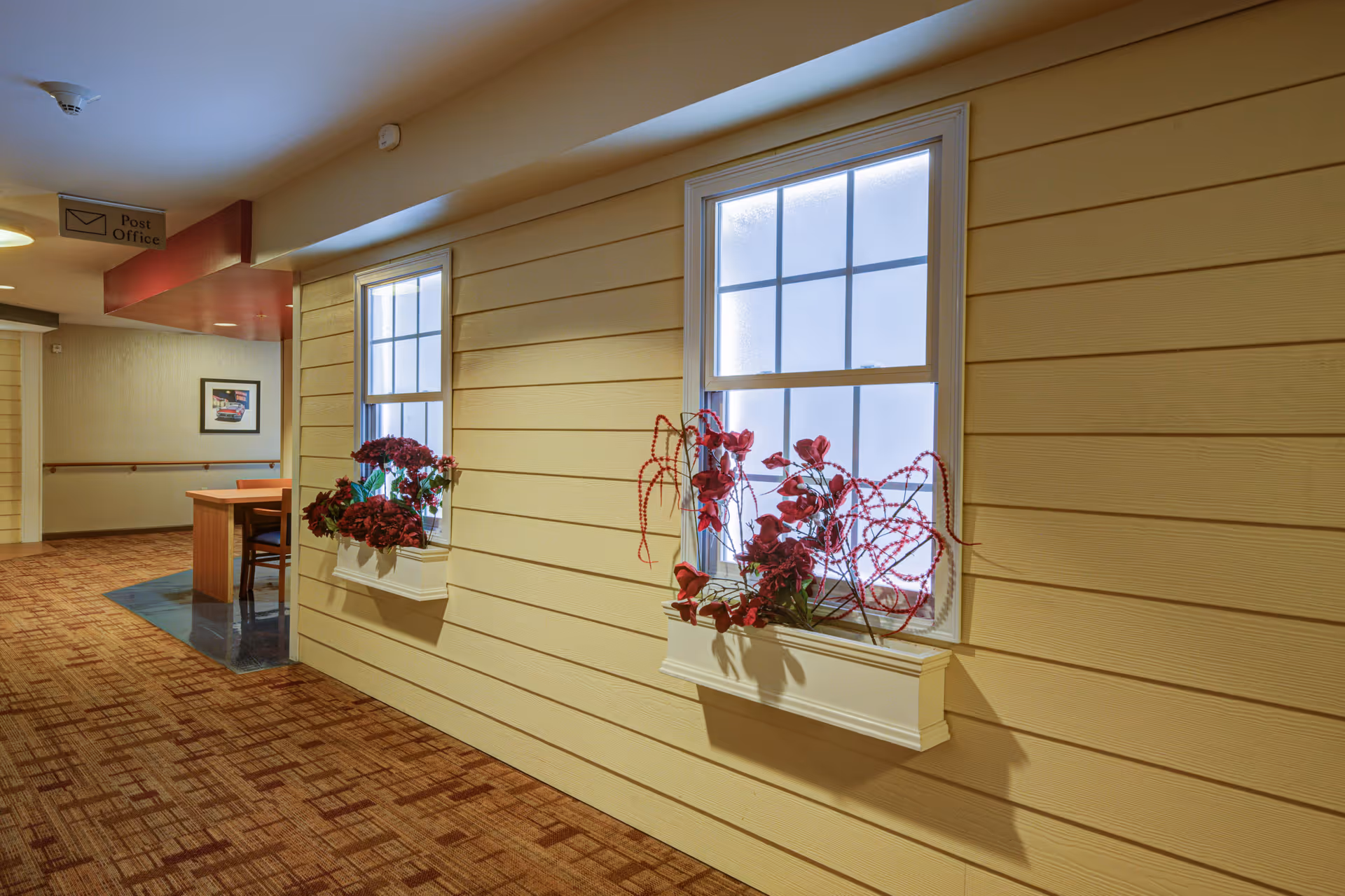 Interior hallway with beige horizontal panel walls and two windows with white frames and flower boxes containing red flowers. The floor is carpeted with a patterned design. In the background, there is a small table with chairs and a sign overhead that reads 'Post Office'.
