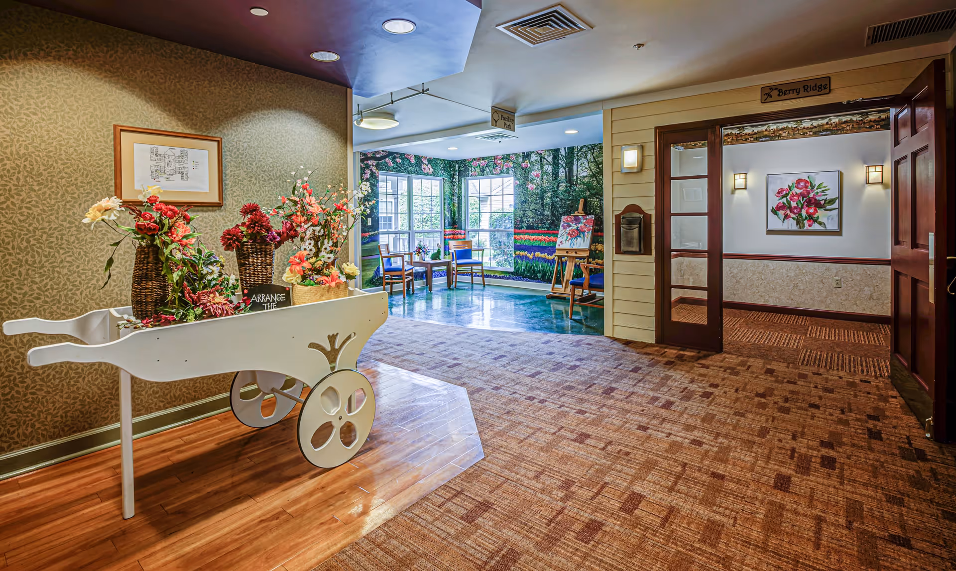 Interior view of a senior living facility hallway with a decorative white cart holding flower arrangements on the left. The hallway has patterned carpet and wood flooring, with a seating area in the background featuring chairs and a mural of a garden with flowers and trees. There is a framed floor plan on the wall and a sign above a doorway labeled 'Berry Ridge'.