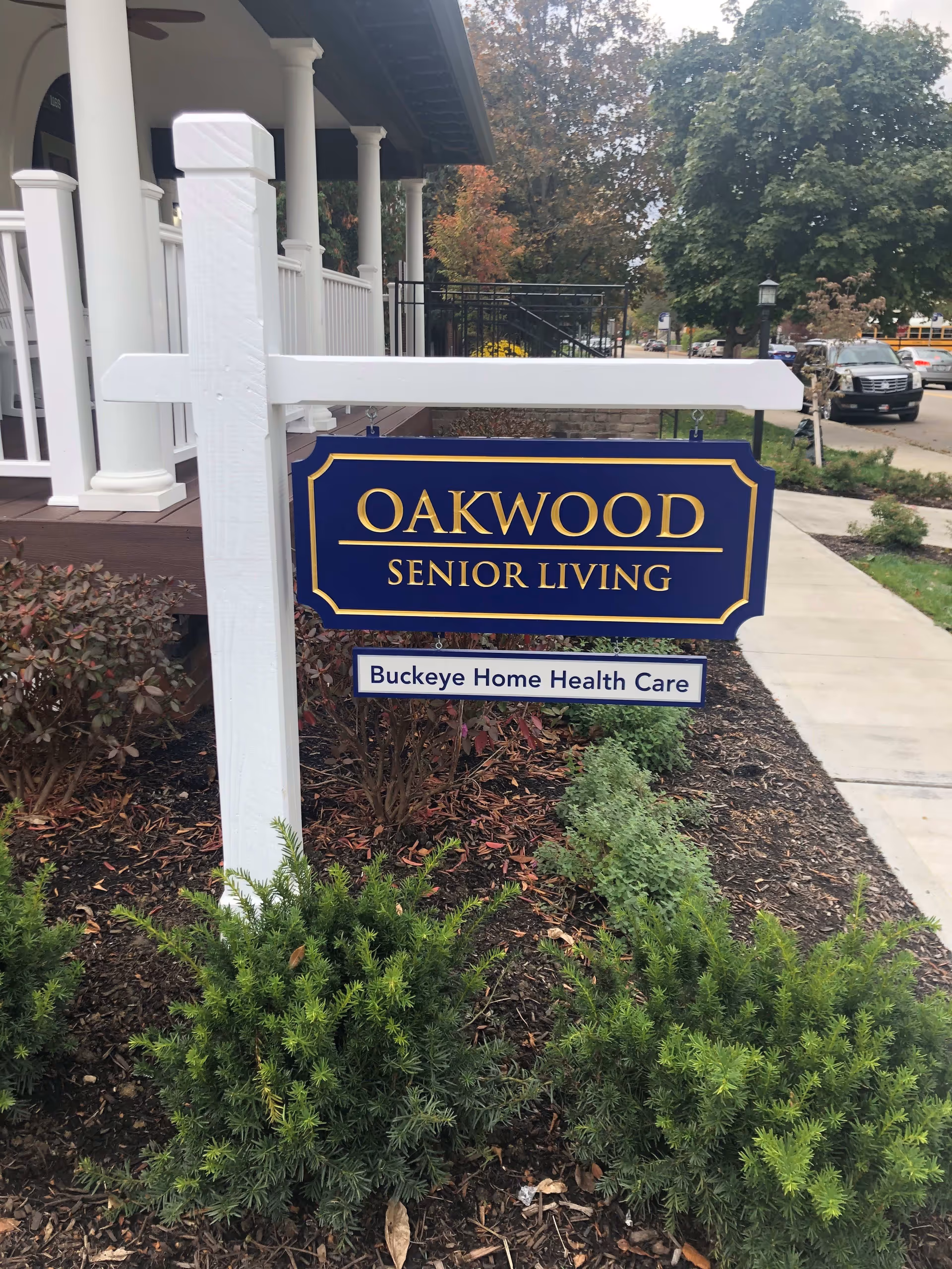 A blue and gold sign reading 'OAKWOOD SENIOR LIVING' with a smaller sign below it that says 'Buckeye Home Health Care' is mounted on a white post in a landscaped area with bushes and mulch. In the background, there is a porch with white columns and a sidewalk leading to a street with parked cars and trees.
