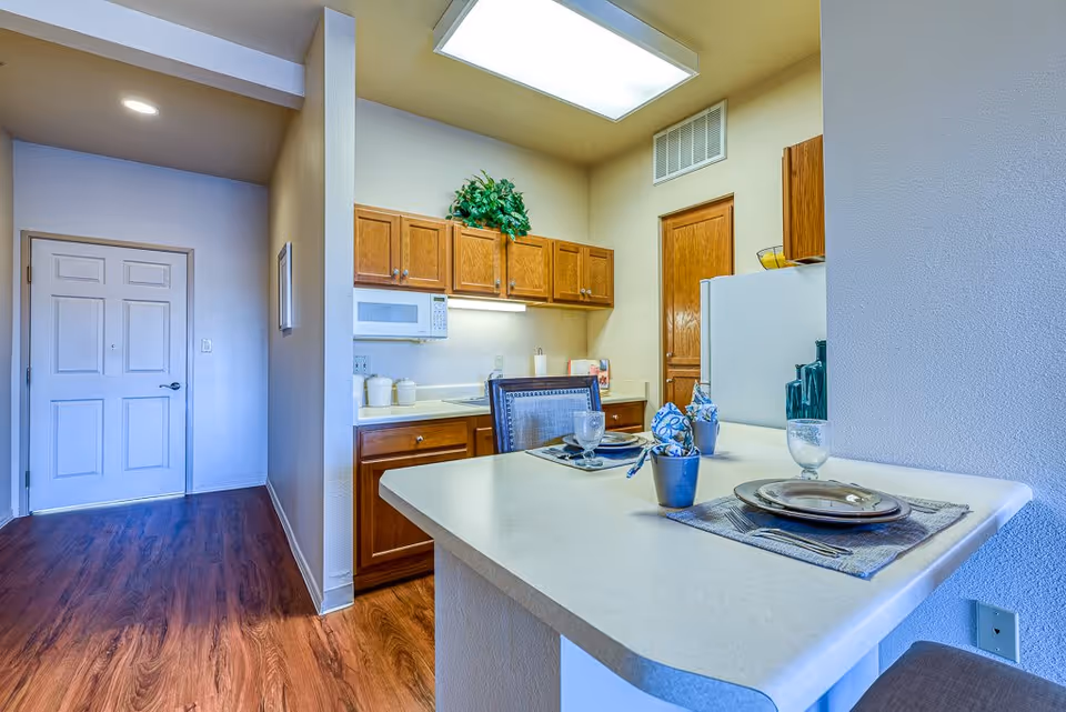Interior view of a kitchen area in a senior living facility with wooden cabinets, a white microwave, a refrigerator, and a countertop set with plates, glasses, and napkins. The floor has wood-like flooring, and there is a white door visible in the background.