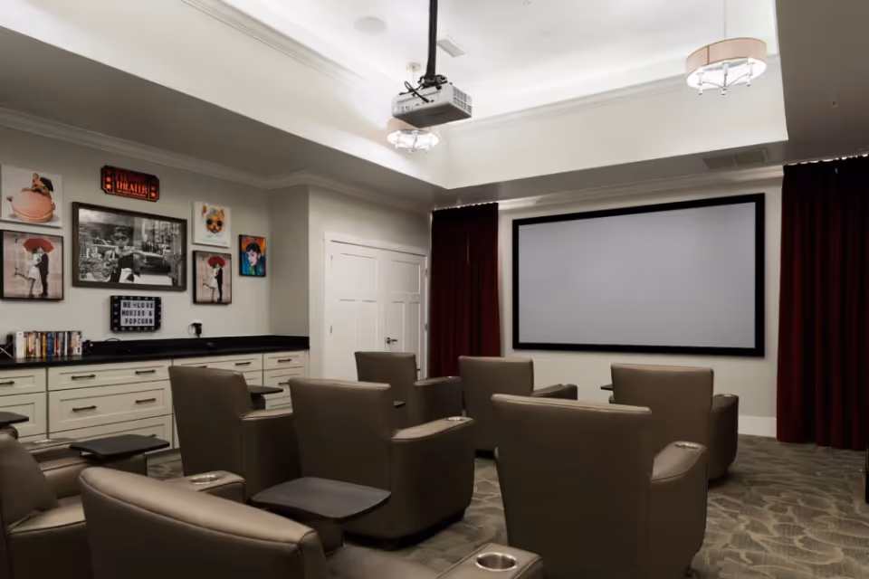 A small home theater room with several brown leather recliner chairs arranged in rows facing a large blank projector screen. The room has a ceiling-mounted projector, dark red curtains on the sides of the screen, and framed movie-themed artwork on the wall above a counter with drawers. The lighting fixtures are modern and the carpet has a subtle pattern.