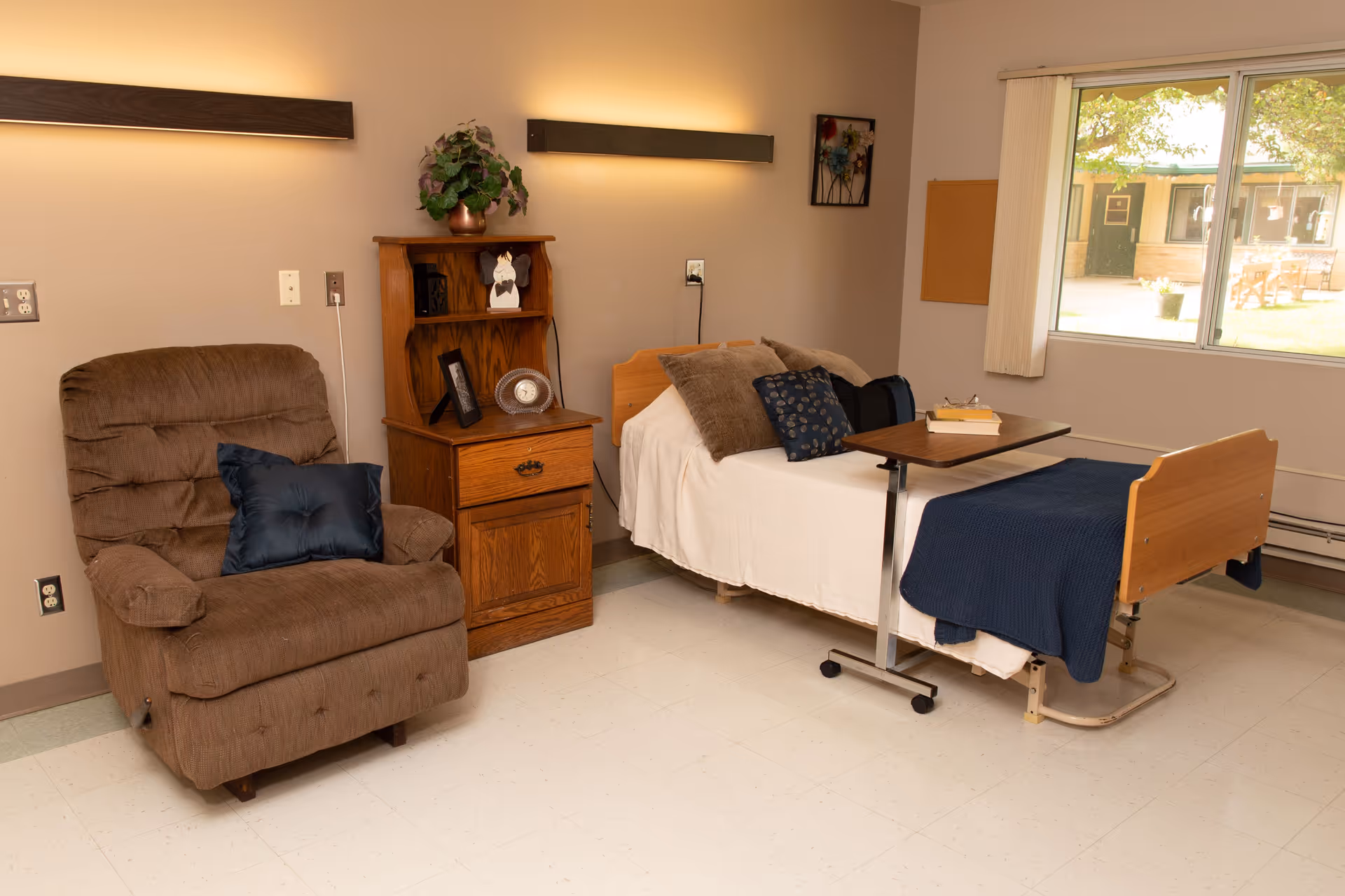 A cozy room with a hospital-style bed covered with white and blue bedding, a brown recliner chair with a blue pillow, a wooden nightstand with decorative items, and a large window showing an outdoor courtyard.