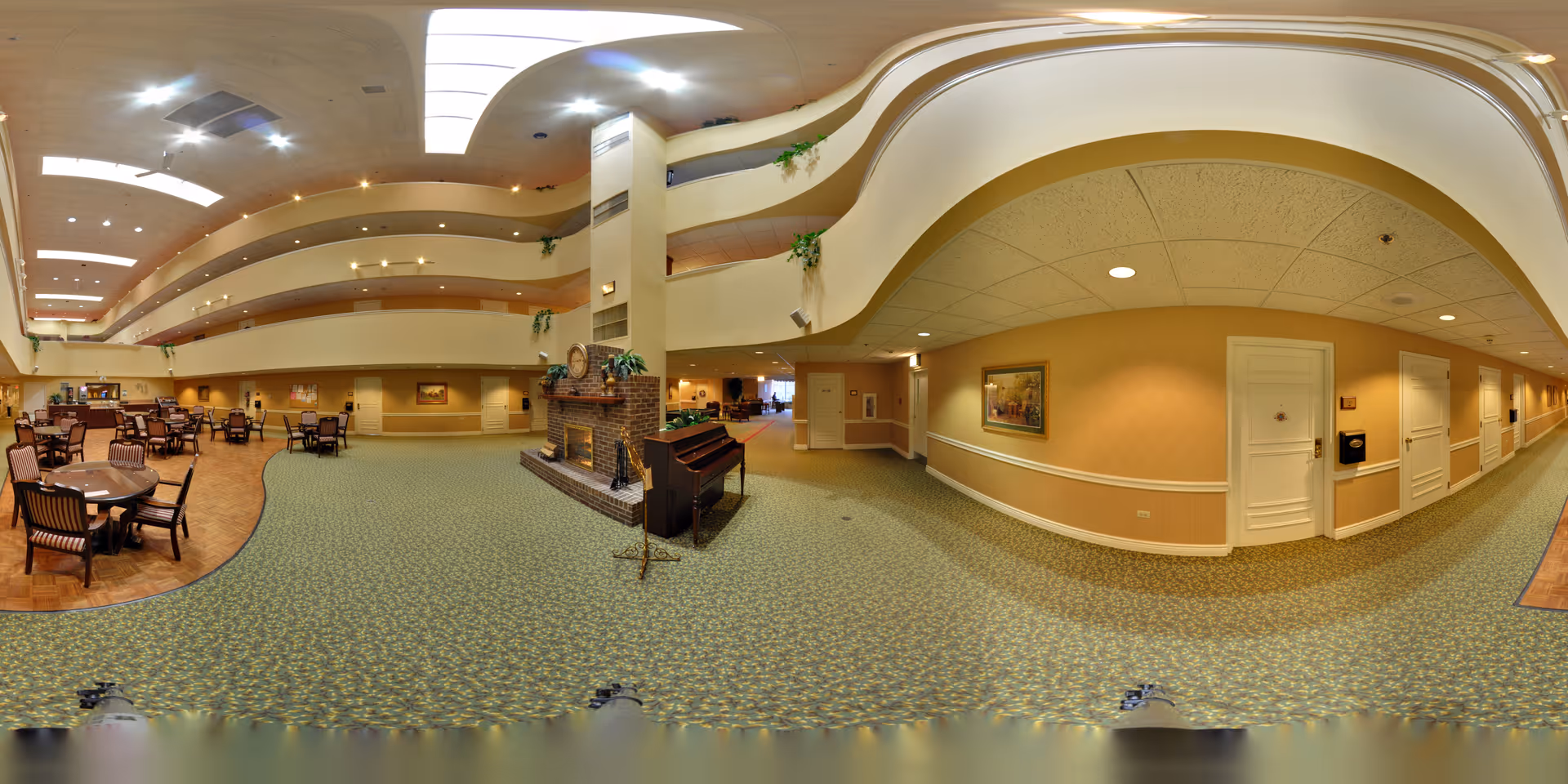 Wide panoramic view of a senior living facility's common area featuring a spacious lounge with multiple tables and chairs on the left side, a carpeted floor, a brick fireplace with a piano in front of it, and a hallway with several doors on the right side. The ceiling has recessed lighting and skylights, and the upper floors with railings are visible above.