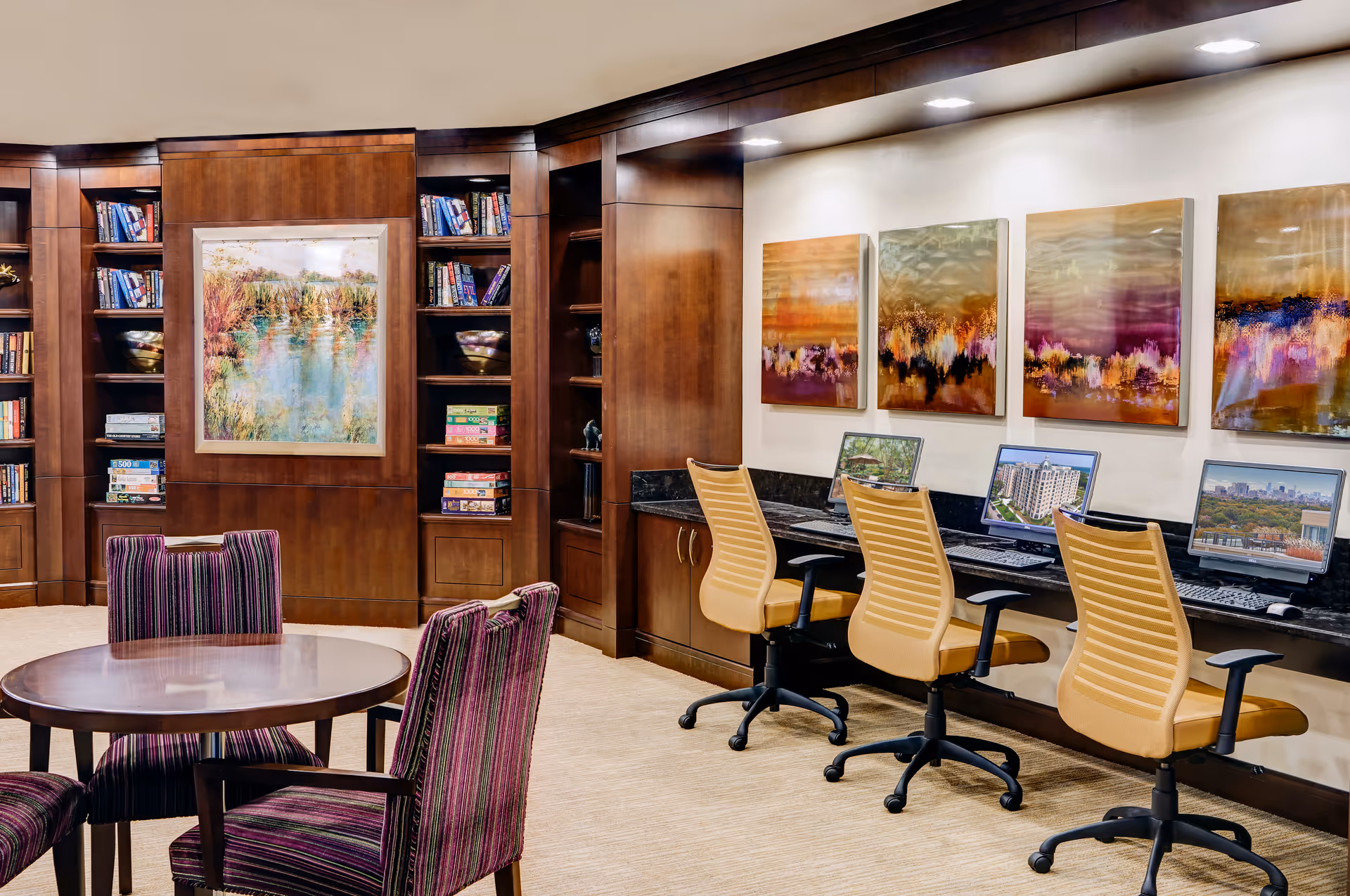A well-lit room with a round wooden table surrounded by four striped upholstered chairs. Along one wall, there are three computer workstations with yellow office chairs and colorful abstract paintings above them. The opposite wall features built-in wooden bookshelves filled with books and board games, and a framed landscape painting is centered between the shelves.