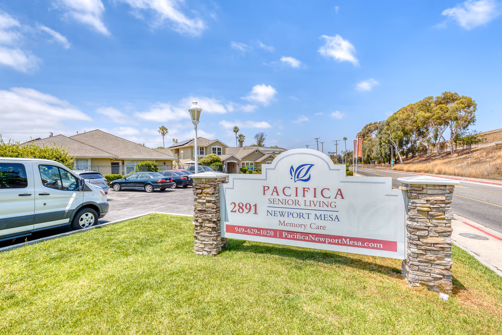 Entrance sign for Pacifica Senior Living Newport Mesa memory care in front of the facility with parked cars and buildings behind.
