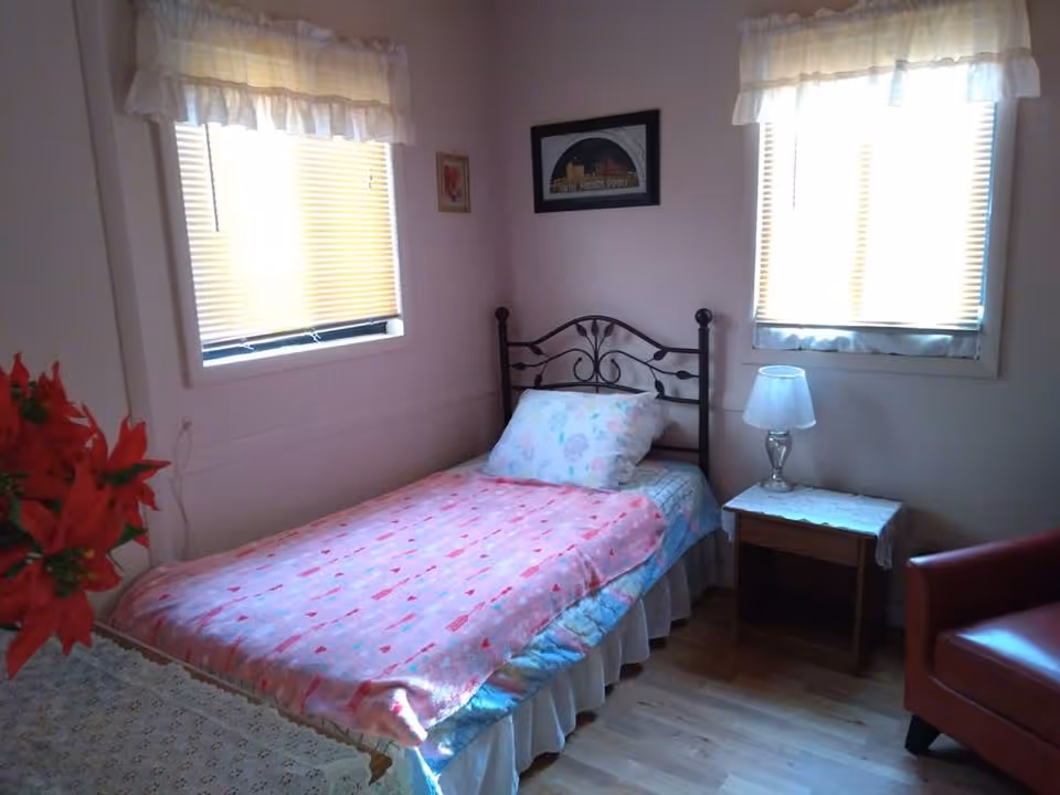 Small single-occupancy bedroom with a metal headboard bed covered in pink bedding, a bedside table with a lamp, two windows, and a red armchair.