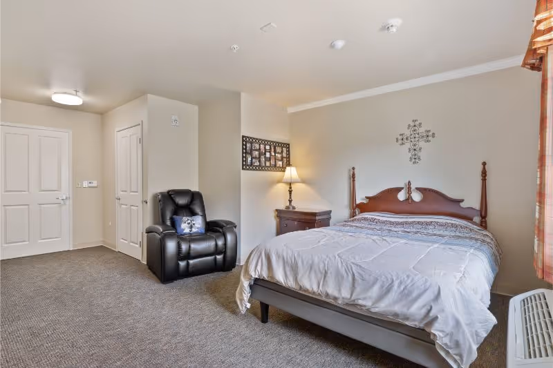 A bedroom in a senior living facility featuring a wooden bed with white bedding, a nightstand with a lamp, a black recliner chair with a decorative pillow, beige walls, carpeted floor, and a window with plaid curtains.