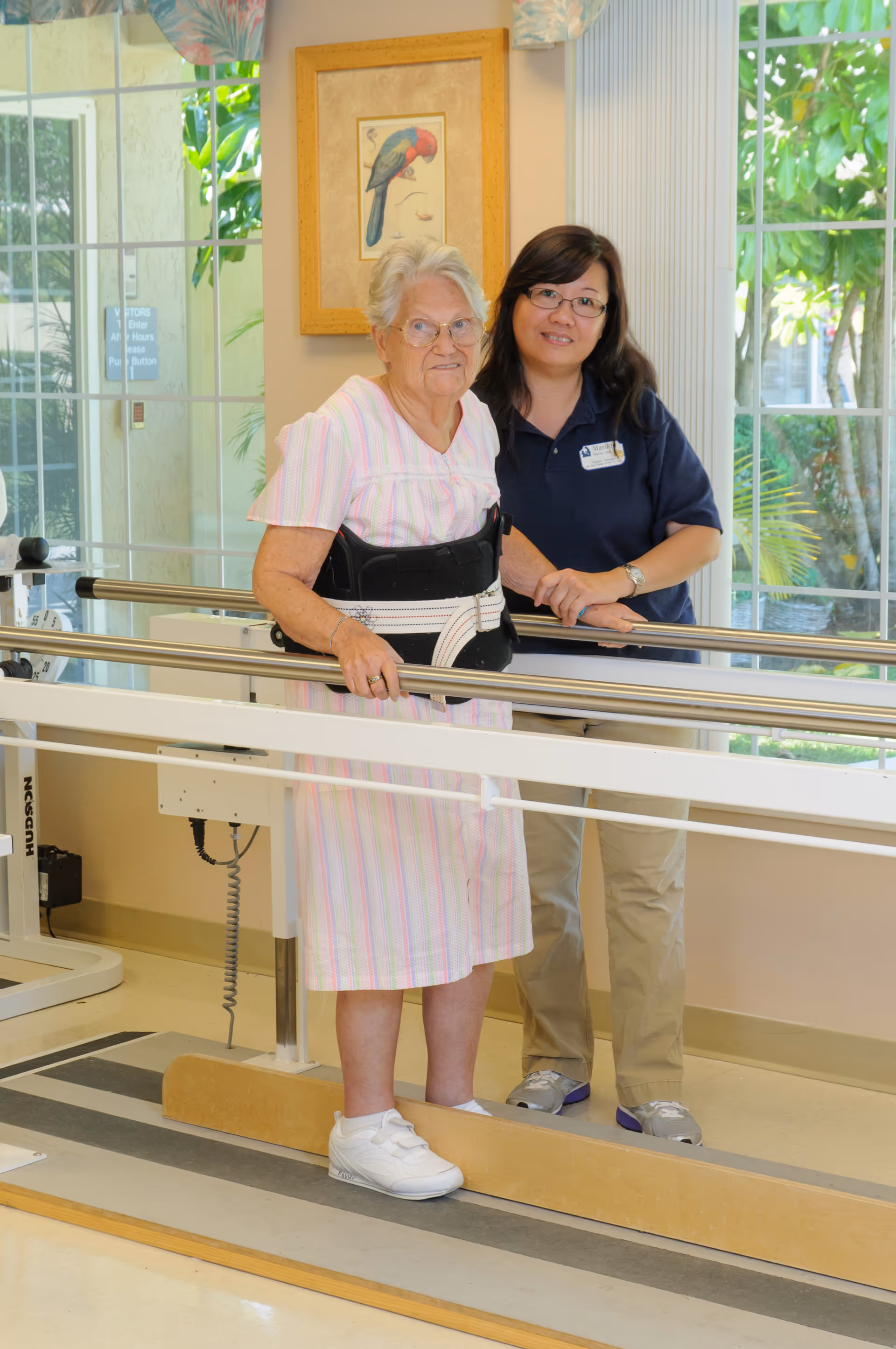 An elderly woman wearing a pink striped dress and a supportive back brace is standing and holding onto parallel bars for physical therapy. A female caregiver or therapist stands beside her, smiling and providing support. The room has large windows with a view of greenery outside and a framed picture of a bird on the wall.