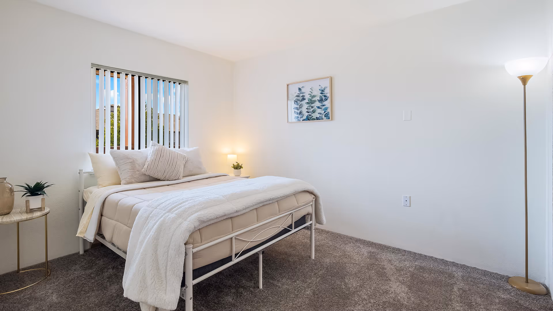 A bright and cozy bedroom with a neatly made bed featuring beige and white bedding and multiple pillows. There is a small round side table with decorative items on the left, a window with vertical blinds letting in natural light, a framed botanical print on the wall, and a tall floor lamp on the right side of the room. The floor is carpeted in a neutral tone.