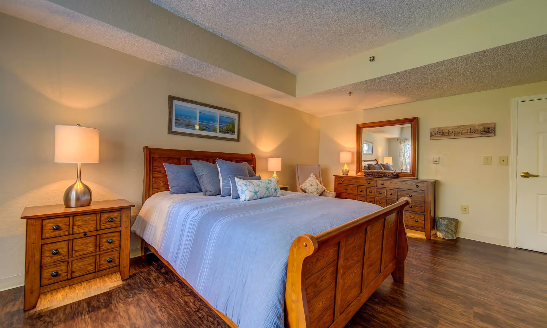 A cozy bedroom featuring a wooden bed with blue bedding, two bedside lamps, a dresser, and a mirror. The room has warm lighting and a calming atmosphere.
