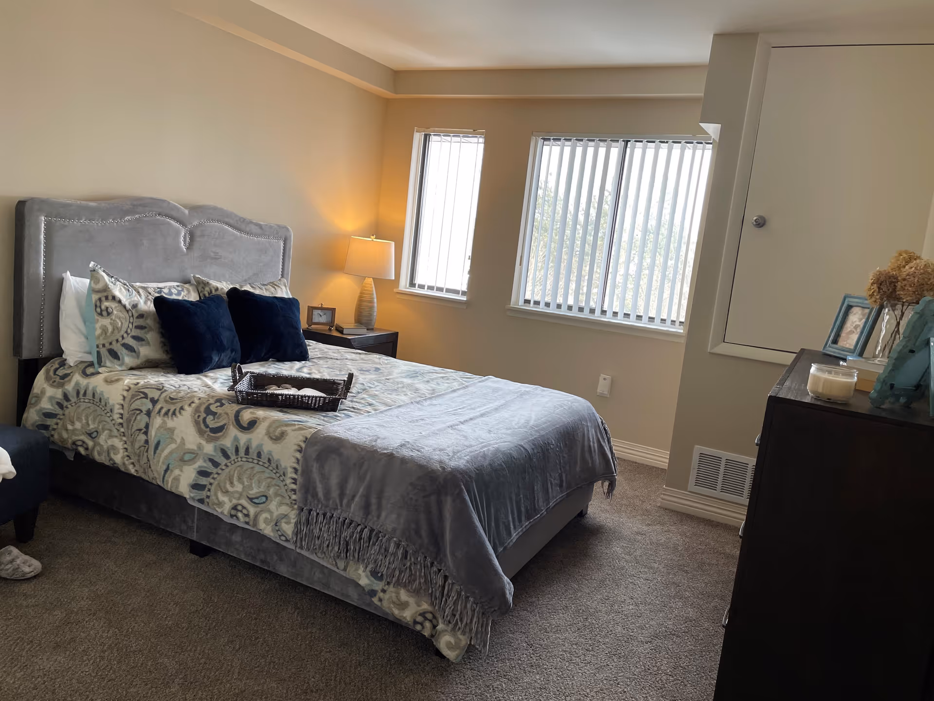 A cozy bedroom with a large bed featuring a gray upholstered headboard, patterned bedding, and two dark blue pillows. There is a nightstand with a lamp and a small picture frame next to the bed. Two windows with vertical blinds allow natural light into the room. A dark dresser with decorative items and picture frames is on the right side of the room. The carpet is gray and the walls are painted a light beige color.